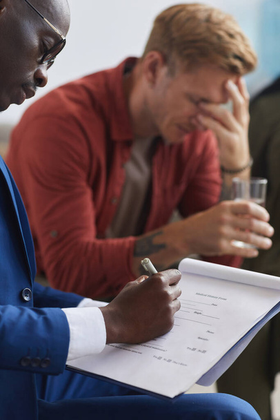 Vista lateral vertical en el psicólogo afroamericano masculino escribiendo en el portapapeles mientras lidera la sesión de grupo de apoyo, espacio para copiar - Foto, Imagen