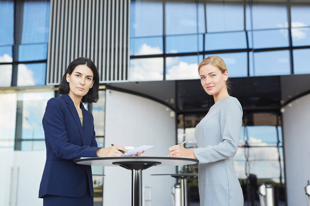 Side view portrait of two businesswomen smiling at camera while standing by cafe table in airport or office building, copy space - Photo, Image