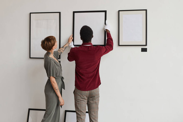 Vista posterior retrato de dos trabajadores de galería de arte colgando marcos de pintura en la pared blanca, mientras que la planificación de la exposición en el museo, espacio de copia - Foto, Imagen