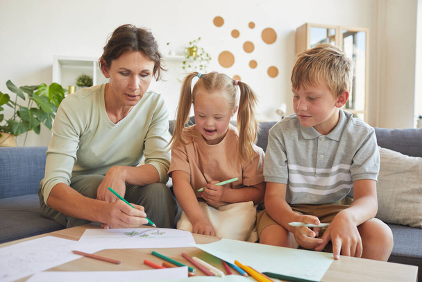 Portrait of cute blonde girl with down syndrome laughing happily while drawing together with mother and brother at home - Foto, Bild