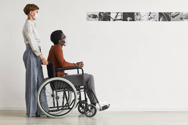 Minimal portrait complet de l'homme afro-américain en fauteuil roulant et en regardant des peintures dans la galerie d'art moderne avec une jeune femme l'aider, les deux portant des masques, espace de copie - Photo, image