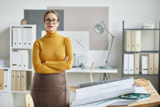 Waist up portrait of confident female architect looking at camera while standing with arms crossed by drawing desk in office, copy space - Foto, Bild