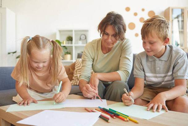 Portrait of cute blonde girl with down syndrome drawing with mother and brother together in home interior - Photo, Image