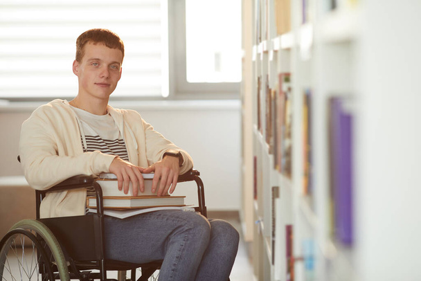 Ritratto caldo di giovane che usa la sedia a rotelle a scuola mentre guarda la macchina fotografica in biblioteca illuminata dalla luce del sole - Foto, immagini