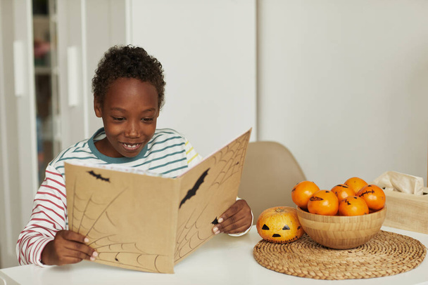 Boldog afro-amerikai fiú ül az asztalnál olvasás érdekes Halloween történet könyv, másolás helyet - Fotó, kép