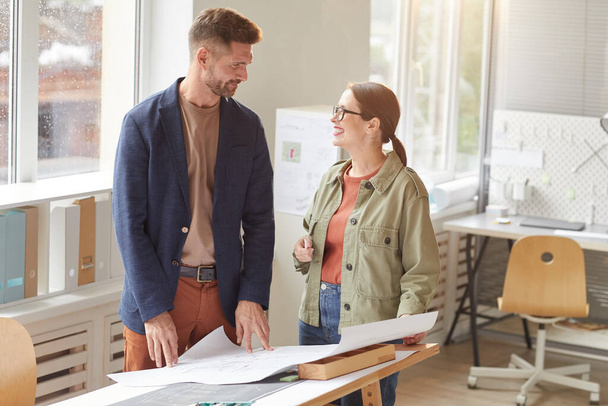 Waist up portrait of two smiling architects discussing blueprints while standing by drawing desk in office , copy space - Foto, imagen