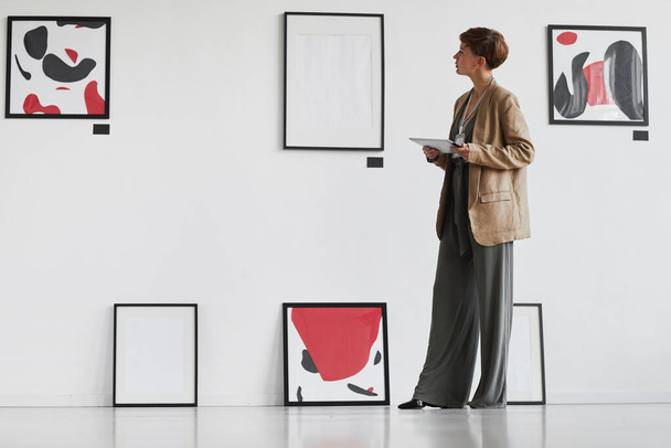 Full length πορτρέτο της δημιουργικής κομψή γυναίκα κοιτάζοντας πίνακες που κρέμονται σε λευκό τοίχο, ενώ επισκέπτονται έκθεση γκαλερί σύγχρονης τέχνης, αντίγραφο χώρου - Φωτογραφία, εικόνα