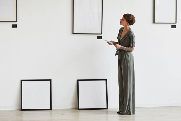 Retrato gráfico de comprimento total de elegante gerente de galeria de arte feminina olhando para a configuração do quadro ao planejar exposição ou evento, espaço de cópia - Foto, Imagem