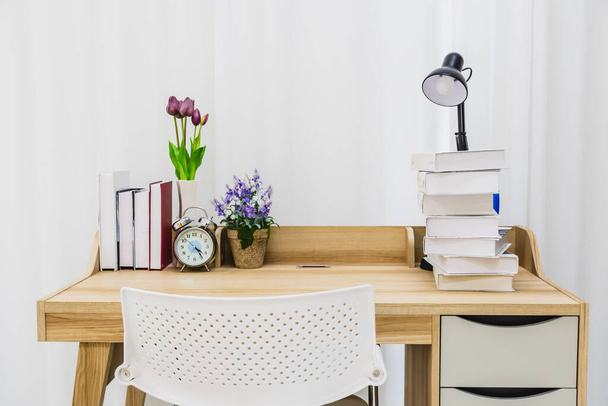 Une table en bois pour lire dans une pièce avec une chaise, une lampe, des livres, une horloge et un pot de fleurs, avec un rideau blanc en arrière-plan. La table du livre pour travailler dans la salle blanche. - Photo, image