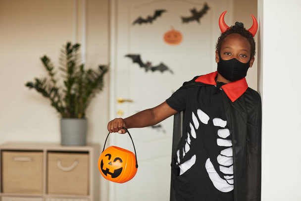 Kırmızı boynuzlu ve maskeli korkunç şeytan kostümü giyen sevimli Afro-Amerikan çocuk portresi kameraya bakan şekerler için fener sepeti tutuyor. - Fotoğraf, Görsel