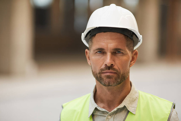 Close-up portret van volwassen bouwvakker dragen van harde hoed en kijken naar de camera tijdens het staan op de bouwplaats, kopieer ruimte - Foto, afbeelding
