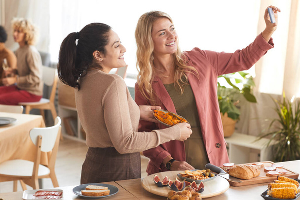 Retrato de dos mujeres adultas modernas tomando foto selfie en el interior mientras disfruta de la cena con amigos, espacio para copiar - Foto, Imagen