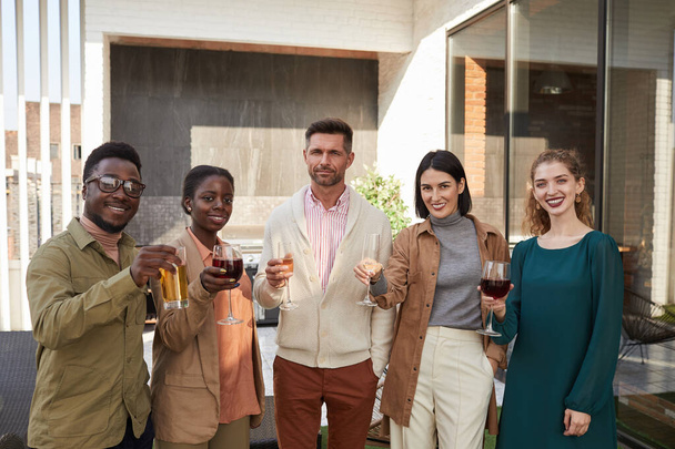 Porträt einer multiethnischen Gruppe von Freunden, die in die Kamera schauen, während sie auf der Außenterrasse während der Party Wein genießen - Foto, Bild