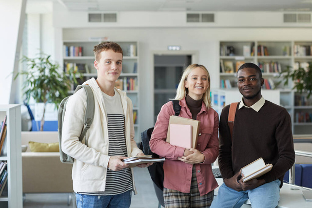 Cintura para cima retrato de três jovens estudantes olhando para a câmera e sorrindo enquanto estava de pé na biblioteca da faculdade - Foto, Imagem