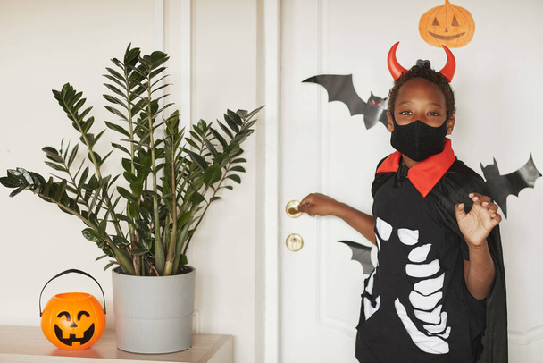 Современный афроамериканец в костюме дьявола с красными рогами и черной маской на лице, прощаясь со своими родителями и отправляясь на сладости с друзьями.. - Фото, изображение