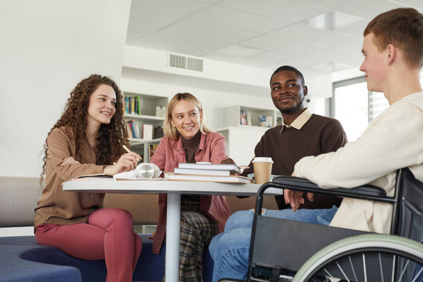 Üniversite kütüphanesinde okuyan çok ırklı bir grup öğrenciye düşük açılı bakış açısı. Önplanda tekerlekli sandalye kullanan bir genç var. - Fotoğraf, Görsel