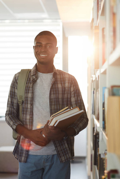 Vertical waist up portrait of young African-American man holding books and smiling at camera while standing in school library - Foto, Bild