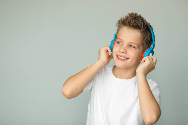 Handsome boy in a white T-shirt listens to music with headphones. The teenager enjoys the music, his eyes are quite glittering. Isolated on a gray background. Large portrait. - Photo, Image