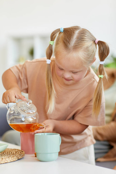 Retrato vertical de menina bonito com síndrome de down poring chá na xícara enquanto desfruta de tempo com a família em casa - Foto, Imagem