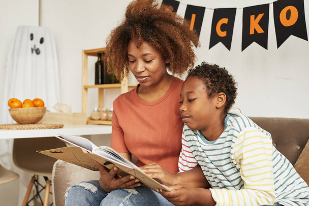 Bela mulher afro-americana sentada no sofá na sala de estar com seu filho pré-adolescente lendo interessante livro de histórias de Halloween - Foto, Imagem