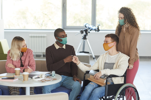Προσωπογραφία πολυεθνικής ομάδας φοιτητών που φορούν μάσκες ενώ σπουδάζουν σε βιβλιοθήκη κολλεγίου με νεαρό άνδρα που χρησιμοποιεί αναπηρικό καροτσάκι σε πρώτο πλάνο, αντιγραφή χώρου - Φωτογραφία, εικόνα