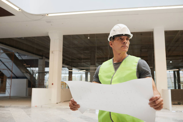 Porträt eines professionellen Bauarbeiters, der Pläne hält und wegschaut, während er im Bürogebäude steht, kopiert Raum - Foto, Bild