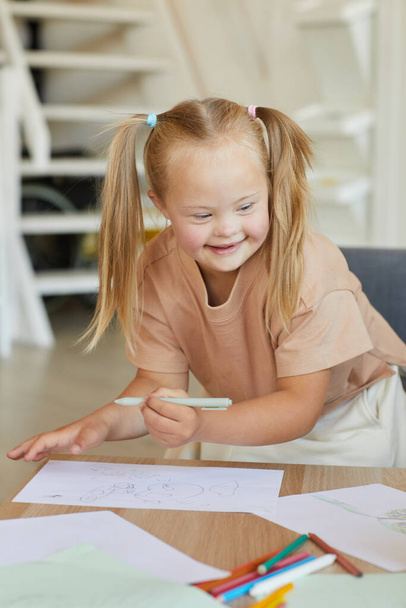 Vertical portrait of cute blonde girl with down syndrome drawing and smiling happily while enjoying development class - Fotó, kép