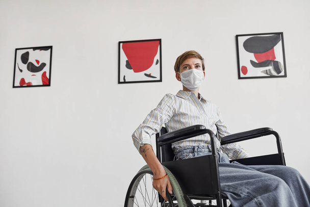 Wide angle portrait of young woman using wheelchair and wearing mask while looking at paintings in modern art gallery - Photo, Image