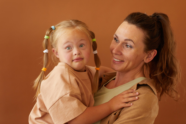 Porträt einer glücklichen Mutter, die ihre Tochter mit Down-Syndrom hält, während sie im Studio vor schlichtem braunen Hintergrund steht - Foto, Bild