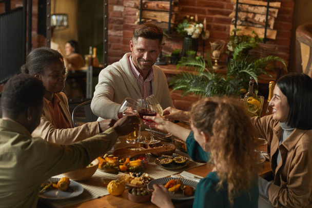 Mnohonárodnostní skupina lidí cinkající sklenice při vychutnávání večeře s přáteli a rodinou v útulném interiéru - Fotografie, Obrázek
