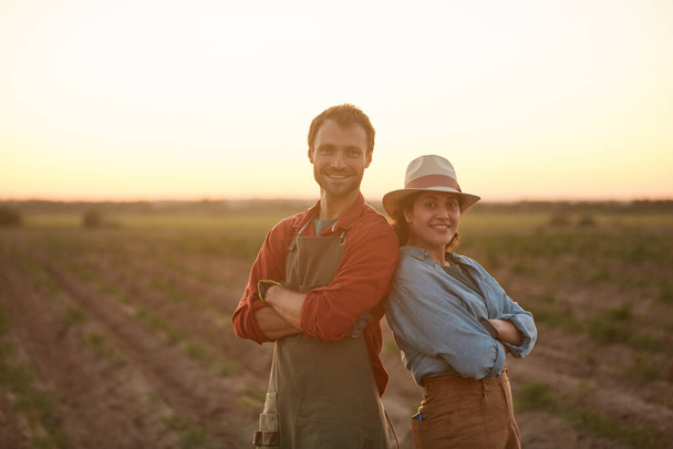 夕暮れ時に畑に立ち、カメラで笑っている間に後ろにポーズをとっている若い農家のカップルの腰を上げ、スペースをコピー - 写真・画像