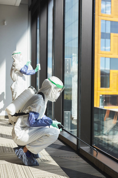 Retrato vertical de dos trabajadores con trajes de materiales peligrosos desinfectando ventanas y pisos en el edificio de oficinas - Foto, imagen