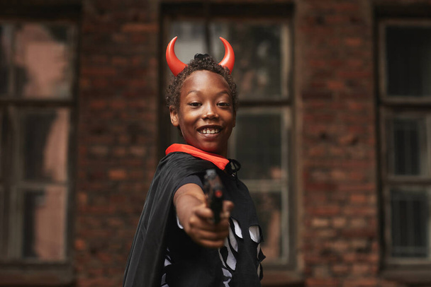 Портрет веселого підлітка, одягненого в диявол з червоними рогами на Хелловін вечірки стоячи проти старого будинку, який стріляв в камері з іграшкового пістолета. - Фото, зображення