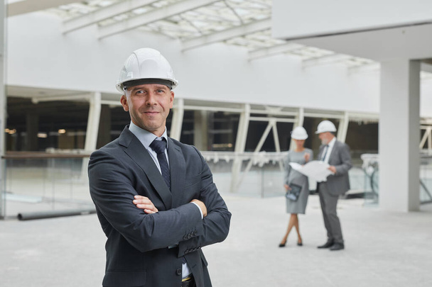 Waist up portrait of smiling mature businessman wearing hardhat and looking at camera while standing with arms crossed at construction site, copy space - Foto, imagen