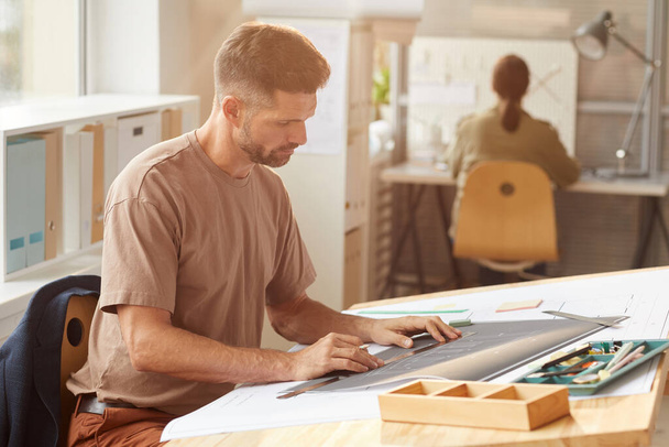 Seitenansicht Porträt eines erwachsenen bärtigen Architekten beim Betrachten von Bauplänen, während er am Zeichentisch im Sonnenlicht sitzt, Kopierraum - Foto, Bild
