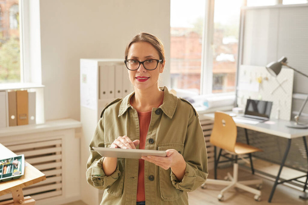 Taille omhoog portret om moderne jonge vrouw met behulp van digitale tablet en kijken naar de camera terwijl het staan in kantoor verlicht door zonlicht, kopieer ruimte - Foto, afbeelding