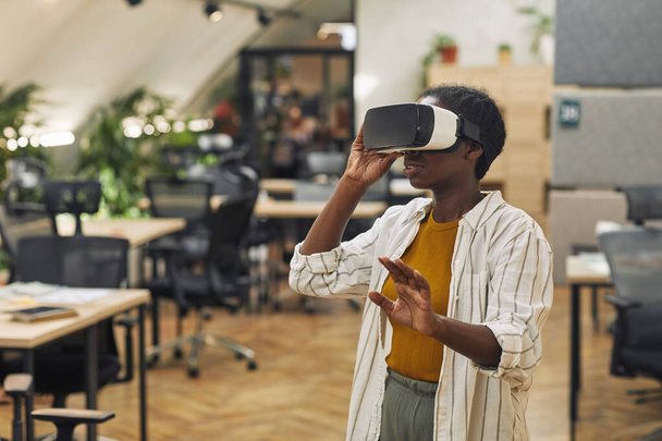 Portrait taille haute de la jeune femme afro-américaine portant un casque VR au bureau et profitant de la réalité immersive, espace de copie - Photo, image