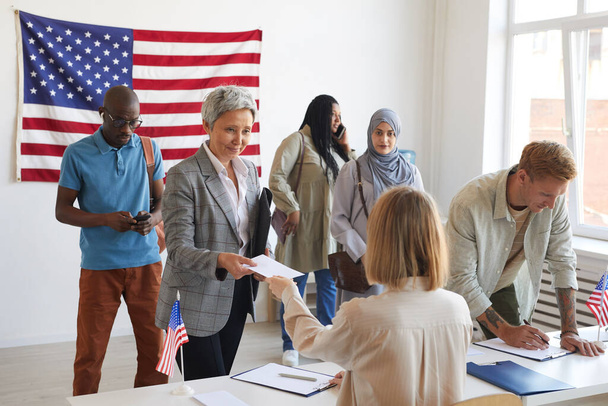 Мультиетнічна група людей реєструється на виборчій дільниці прикрашена американськими прапорами в день виборів, копіюйте простір. - Фото, зображення