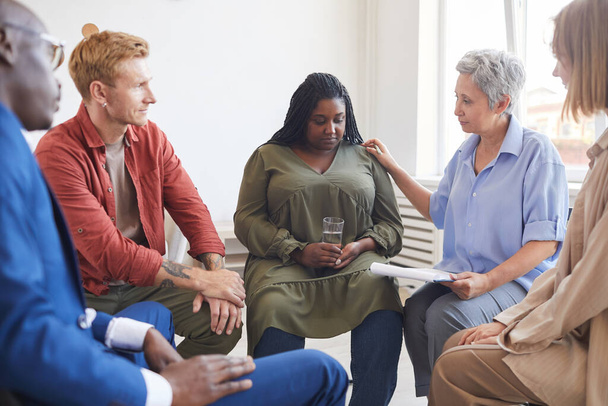 Portrait of young African-American woman sharing struggles during support group meeting with people siting in circle and comforting her - Photo, Image