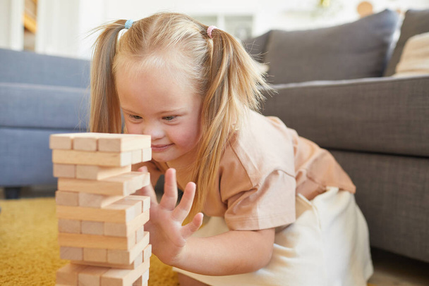 Portrait of cute blonde girl with down syndrome playing board games stacking wood blocks while sitting on floor at home, copy space - Photo, Image