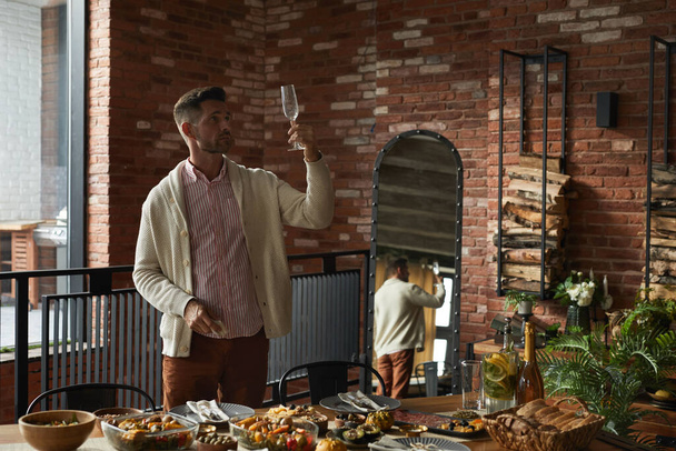 Retrato de un elegante hombre maduro inspeccionando vasos mientras sirve la mesa de cena para la fiesta de Acción de Gracias en casa, espacio para copiar - Foto, imagen
