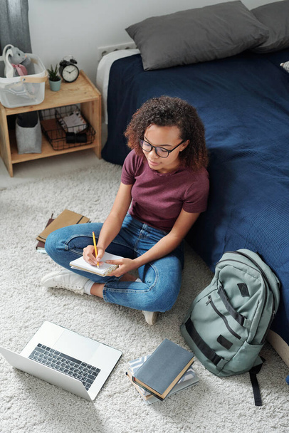 Genç, çapraz bacaklı bir öğrenci dizüstü bilgisayarın önünde oturuyor ve online ders sırasında öğretmenini dinlerken notlar alıyor. - Fotoğraf, Görsel