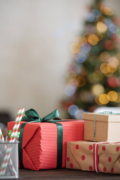 Grupo de regalos de Navidad embalados envueltos en papel y atados con cintas en la mesa frente a la cámara contra el árbol de Navidad decorado - Foto, imagen