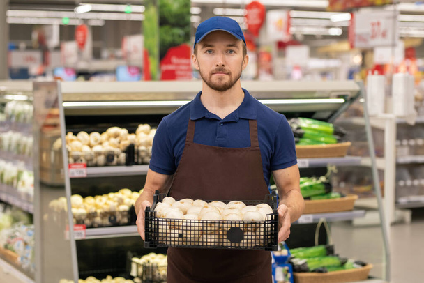 Vertikale mittlere Langzeitaufnahme eines modernen Ladenarbeiters in Uniform, der eine Box voller frischer Pilze trägt - Foto, Bild
