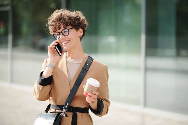 Young cheerful businesswoman with drink in hand and smartphone by ear consulting client outdoors against building exterior on sunny day - Photo, Image