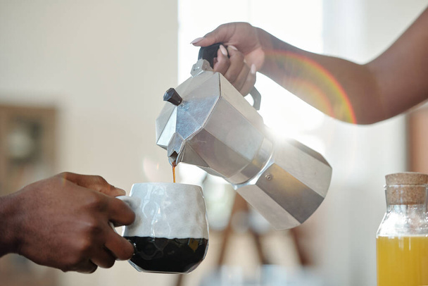 Main de jeune femme africaine versant du café ou du thé dans la tasse de son mari pendant le petit déjeuner avec bouteille de jus d'orange à proximité - Photo, image
