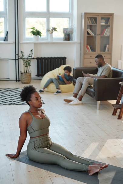 Fit jeune femme africaine en survêtement gris assis sur le tapis sur le sol pendant l'exercice physique contre son mari et son petit fils reposant sur le canapé - Photo, image