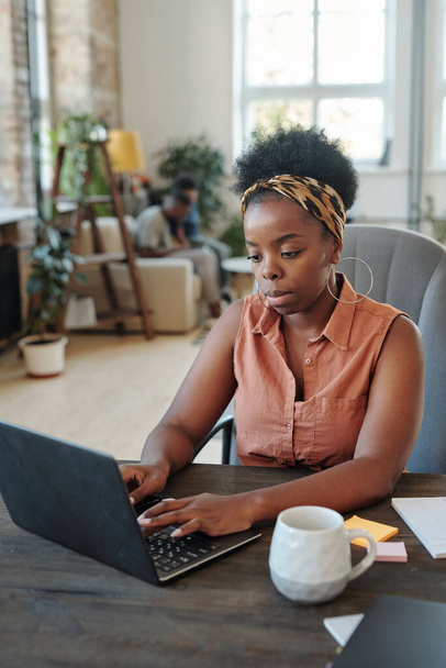 Σοβαρή νεαρή Αφρικανή freelancer σε καφέ casual μπλούζα και λεοπάρ εκτύπωση headband δικτύωσης μπροστά από το laptop στο σπίτι - Φωτογραφία, εικόνα