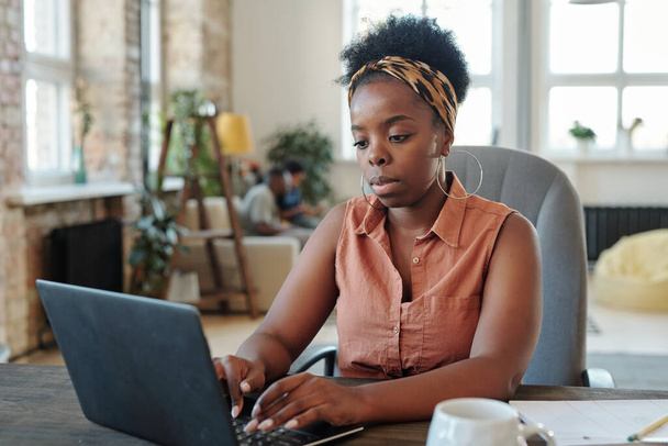 Ev ortamında kucak kucağa otururken dizüstü bilgisayar başında çalışan ciddi Afrikalı serbest çalışan genç bir bayan. - Fotoğraf, Görsel