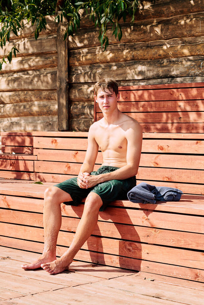 Mladý mokrý muž bez košile surfař v zelených kraťasech sedí na dřevěné lavičce před kamerou v slunečný den po tvrdém venkovním tréninku - Fotografie, Obrázek